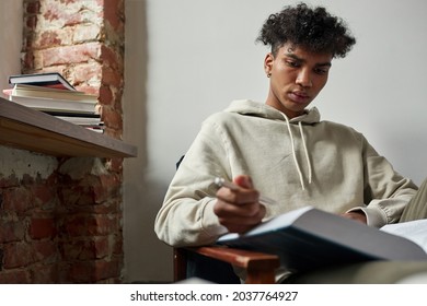 Millennial Gen Z African American Man Sit At Chair At Home Make Notes Think Read Book. Pensive Serious Young Teen Mixed Race Ethnicity Male Study With Textbook. Diversity, Distant Education Concept.