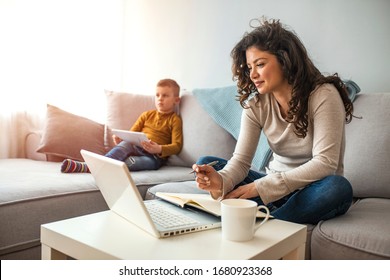 Millennial generation mother working from home with small children while in quarantine isolation during the Covid-19 health crisis. Little boy on tablet computer. Horizontal indoors waist up shot 