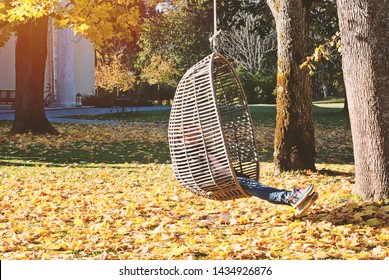 Millennial girl in autumn park swinging on hanging rattan egg-shaped chair outdoor in the midst of fallen leaves