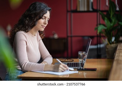 Millennial Girl Sit At Desk Study On Laptop Watch Financial Webinar Making Notes. Concentrated Small Business Owner Work On Computer Write In Notebook, Take Online Course, Training At Home, Education