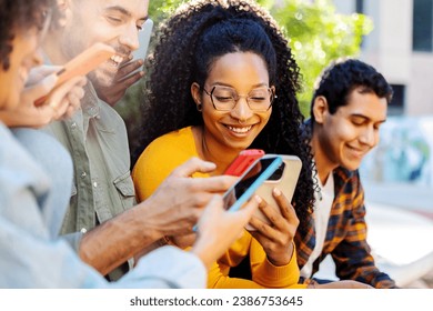 Millennial group of students using mobile phones together at campus college. African american woman having fun watching social media content on cellphone app sitting with her friends outdoors.