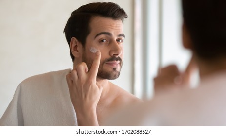 Millennial Handsome Man Looking In Mirror, Applying Moisturizing After Shaving Cream On Cheeks In Bathroom, Head Shot Close Up. Well Groomed Young Guy Doing Skincare Morning Routine After Showering.