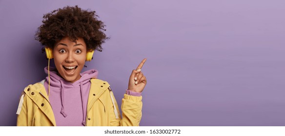 Millennial hipster girl with Afro hair enjoys music podcast in headphones, points fore finger on copy space for your advertising, has excited surprised happy expression, isolated on purple background - Powered by Shutterstock