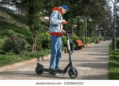 Millennial hipster guy activating electric scooter from smartphone in park. Teenage boy using mobile application to rent e-scooter, standing in city street. People and eco green transportation - Powered by Shutterstock