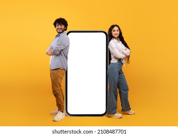 Millennial indian couple leaning on giant smartphone with mockup, promoting mobile application or website, advertising your service or product over yellow studio background