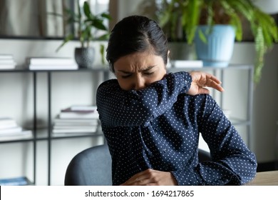 Millennial indian ethnic girl sitting in office, coughing in elbow, right illness behavior for not spreading virus infection. Unhealthy young woman feeling unwell at workplace, covid 19 symptoms. - Shutterstock ID 1694217865