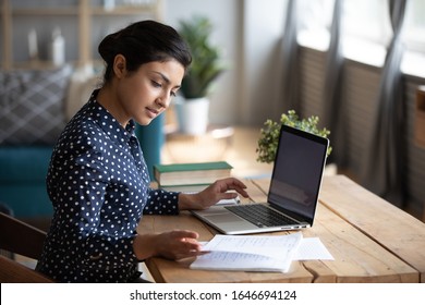 Millennial Indian girl sit at desk in living room study on laptop making notes, concentrated young woman work on computer write in notebook, take online course or training at home, education concept - Shutterstock ID 1646694124