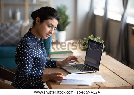 Photo of Millennial Indian girl sit at desk in living room study on laptop making notes, concentrated young woman work on computer write in notebook, take online course or training at home, education concept
