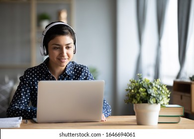 Millennial Indian Girl In Wireless Headphones Sit At Desk At Home Working On Modern Laptop, Young Ethnic Woman In Earphones Browsing Internet Shopping Online Or Studying On Computer In Living Room