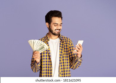 Millennial Indian Guy With Cash Making Money Online On Mobile Phone, Lilac Background