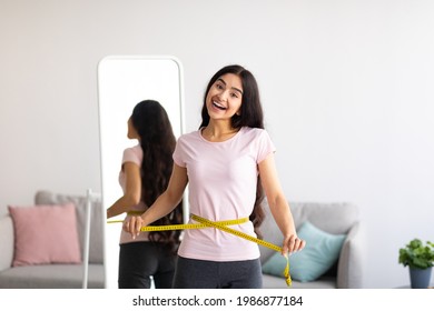 Millennial Indian woman measuring her waist with tape in front of mirror, showing results of slimming diet or liposuction at home. Young Asian lady promoting healthy eating for weight loss