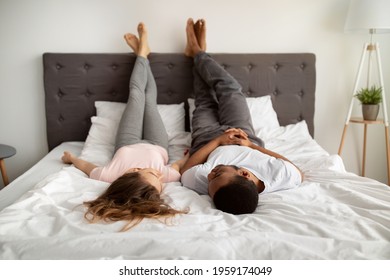 Millennial interracial couple in love lying on bed at home, raising legs up, talking to each other. Lazy young family resting on weekend, communicating, having fun together indoors