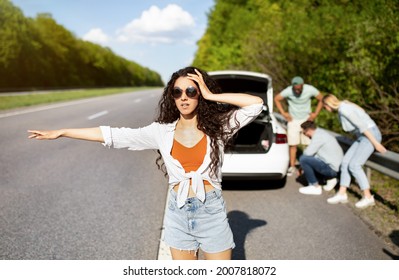 Millennial lady trying to stop passing car, asking for help, her friends fixing broken vehicle, having road accident. Young woman hitchhiking on highway, needing emergency assistance - Powered by Shutterstock