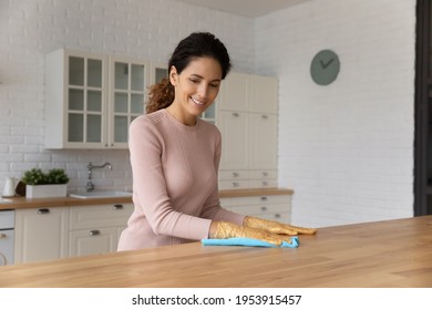 Millennial latin housewife enjoy easy putting modern designed kitchen in order wipe dust from wooden counter. Professional cleaning lady in rubber gloves remove dirt from work top using soft duster