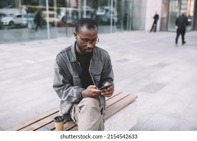 Millennial Male Blogger In Eyeglasses Checking Received Booking Confirmation On Mobile Technology, African American Hipster Guy Browsing Wireless Web Page On Cellular Device Using 4g Internet In City