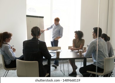 Millennial Male Coach Or Trainer Make Flipchart Presentation For Multiracial Work Group In Office, Man Speaker Or Presenter Draw Discuss Project With Diverse Colleagues At Briefing In Conference Room