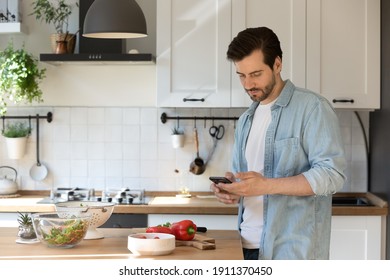 Millennial Male Distracted From Preparing Food Hold Smartphone Order Missing Ingredients Online. Young Man Husband Stand At Modern Kitchen Using Cell Consulting With Wife About Cooking Process Details