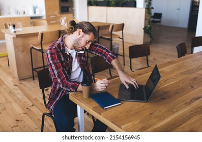 Millennial male software developer browsing laptop website while calling to freelance colleague for discussing marketing details, skilled man with digital netbook making consultancy conversation