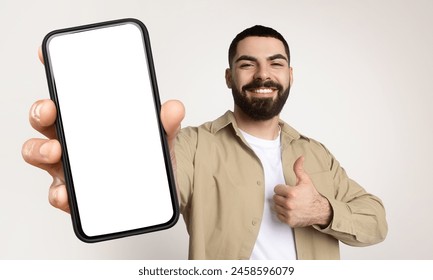 Millennial man is holding a smart phone with white blank screen in his hand, showing thumb up, isolated on beige studio background
