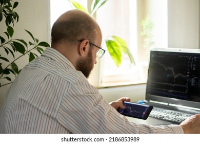 Millennial Man In Light Shirt With Mobile Phone In Hand. Businessman With Glasses Using Mobile Phone, Working From Home,  Looking At Multiple Screens Monitoring Stock