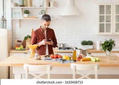 Millennial Man Preparing Romantic Dinner On Home Kitchen. Serious Concentrated Guy Looking For Good Recipe At Smart Phone. Healthy Food Menu App. Cookbook Mobile Application. Technology And Home