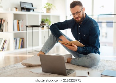 Millennial Man Reading A Book Near Laptop Computer Learning Online Sitting On Floor At Home, Wearing Eyeglasses. Business Literature And Weekend Leisure, Career And Self Growth Concept