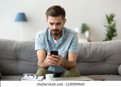 Millennial Man Sit On Couch Using Smartphone Shocked By Reading Online News, Surprised Male Get Breaking Message Or Text On Mobile Phone, Confused Guy Holding Cell See Virus Warning Notification