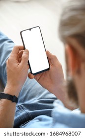 Millennial Man User Holding Smart Phone Mock Up White Screen In Hand Using Mobile Dating App, Check Social Media, Order Food Delivery, Reading News Sit On Couch At Home. Over Shoulder Vertical View.