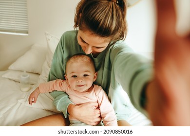 Millennial Mom Taking A Selfie With Her Adorable Baby. Happy Single Mom Taking A Picture While Sitting With Her Newborn Baby On The Bed. New Mom Creating Cute Memories With Her Infant Child.