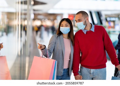 Millennial Multiracial Couple In Face Masks Shopping At Supermarket, Pointing At Store Window, Choosing Goods. Young Diverse Family With Gift Bags Buying Stuff At Mall During Coronavirus Pandemic