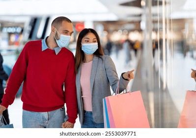 Millennial multiracial couple in face masks shopping at supermarket, pointing at store window, choosing goods. Young diverse family with gift bags buying stuff at mall during coronavirus pandemic - Powered by Shutterstock