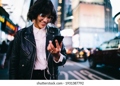 Millennial party blogger sharing publication with content information about nightlife in illuminated New York, prosperous influencer in electronic headphones watching streams video on mobile gadget