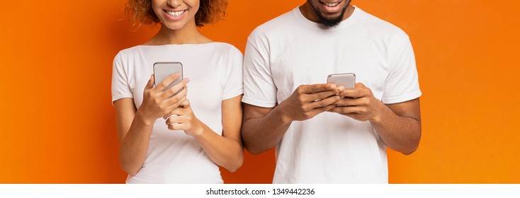 Millennial People Communication. Black Girl And Guy Texting On Mobile Phones, Standing Side By Side On Orange Background, Panorama