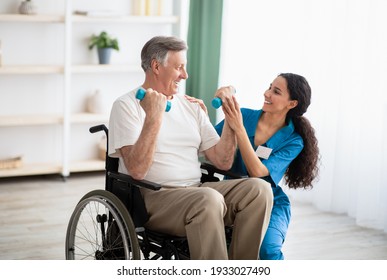 Millennial Physiotherapist Helping Disabled Elderly Man In Wheelchair To Work Out With Dumbbells, To Recover From Injury At Health Centre. Rehab Fitness For Senior Patients Concept