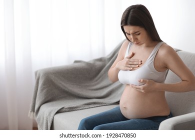 Millennial pregnant lady having painful feelings in breast area, suffering discomfort, young expectant woman massaging her chest while sitting on couch at home, free space
