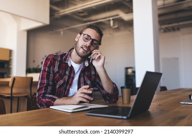 Millennial student with netbook computer and education textbook calling via cellular app for discussing homework while e learning, skilled programmer in spectacles have phone consultancy conversation