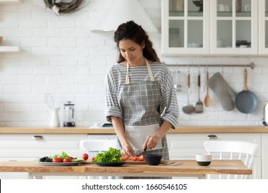 Millennial woman in apron stand at kitchen table chop vegetables preparing salad for dinner or lunch, young housewife cooking healthy tasty breakfast at home, dieting, vegetarian concept