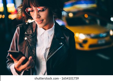 Millennial Woman In Classic Glasses With Neon Reflection Reading Received Notification For Media Application On Modern Cellular Technology, Attractive Generation Z In Trendy Outfit Using Smartphone