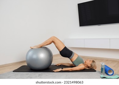 Millennial woman exercising with fitness ball on exercise mat in living room. Fitness, sport and healthy lifestyle concept.