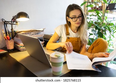 Millennial woman gardener in yellow overalls using laptop, examines project documentation, makes a report on paper documents, works from home. Cozy office workplace, remote work, distant education.  