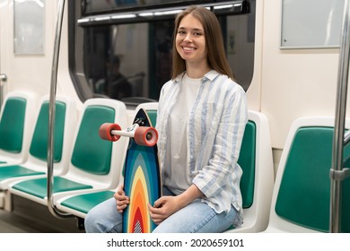 Millennial woman with long hair holding longboard sitting in subway train, smiling, looking at camera. Portrait of happy European young girl skateboarder with skate board in metro train. Urban hobby.