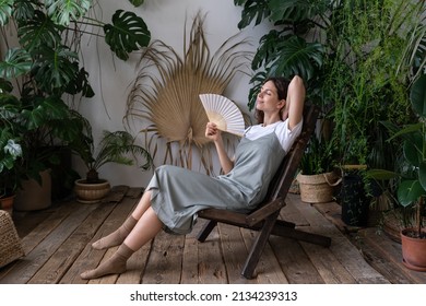 Millennial Woman Rest In Armchair With Closed Eyes Enjoy Fresh Air Hold Fan Cooling Herself At Summer Hot Day In Indoor Garden. Serene Young Female Relax At Home. Comfort Wellbeing Lifestyle Concept