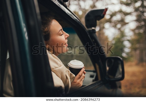 Millennial woman\
travelling off-road by car in a pine forest, drinking coffee,\
enjoying wind. Wanderlust\
concept.