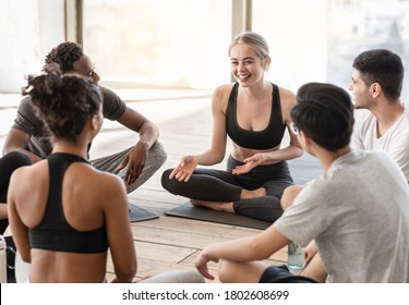 Milti-ethnic group of sporty people chatting and laughing before training in studio, discussing something while sitting on mats at floor, free space