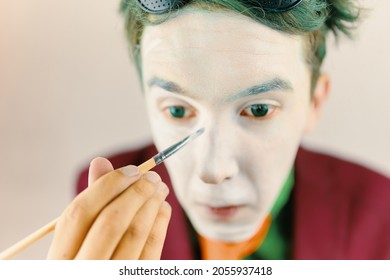 Mim is preparing makeup for performance. Clown cosplayer concept. Man with green hair in burgundy suit. Theatre atmosphere. Male actor is covering his face by white greasepaint using brush.