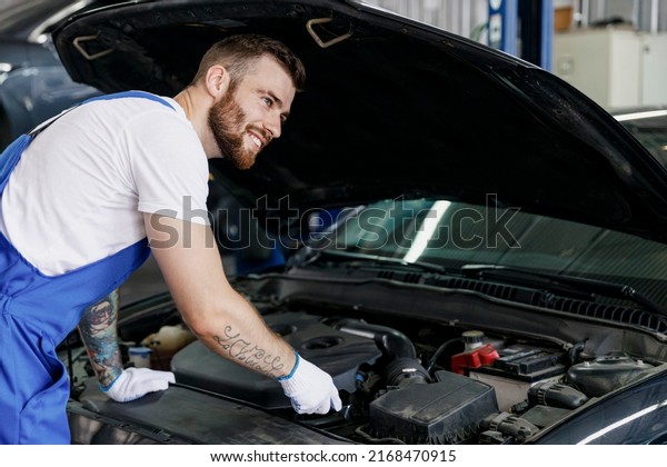 Minded male professional technician car mechanic man\
in denim blue overalls white t-shirt fixing problem with raised\
hood work in light modern vehicle repair shop workshop indoor.\
Tattoo translate fun