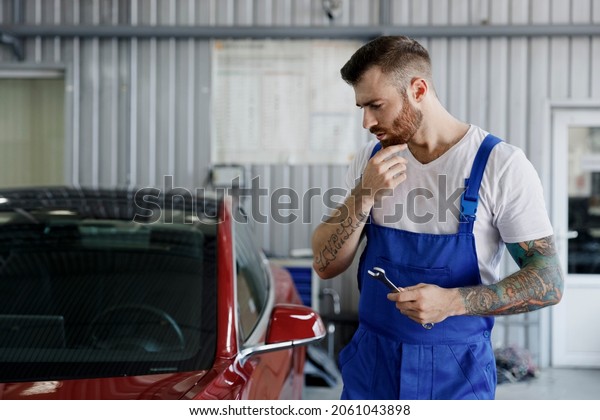 Minded puzzled young male professional\
technician car mechanic repairman man 20s wear denim blue overalls\
white t-shirt hold wrench key tool work in light modern vehicle\
repair shop workshop\
indoors.