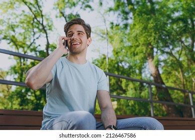 Minded young happy smiling man 20s wearing blue t-shirt sit on bench talk speak on mobile cell phone look aside rest relax in sunshine spring green city park outdoor on nature Urban leisure concept - Powered by Shutterstock