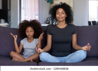 Mindful african mom with cute funny kid daughter doing yoga exercise at home, calm black mother and mixed race little girl sitting in lotus pose on couch together, mum teaching child to meditate