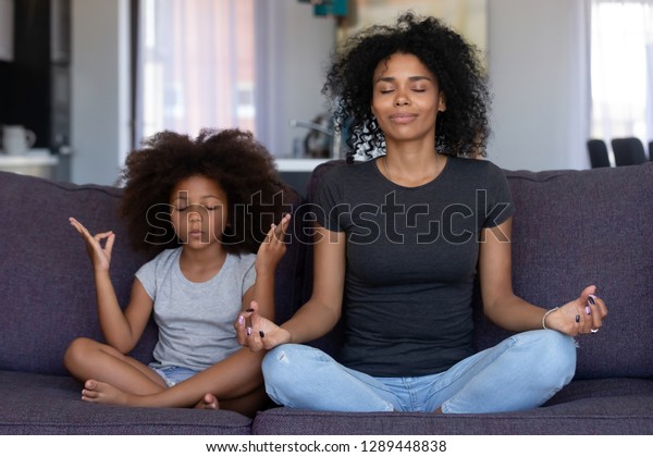 Mindful african mom with cute funny kid daughter\
doing yoga exercise at home, calm black mother and mixed race\
little girl sitting in lotus pose on couch together, mum teaching\
child to meditate